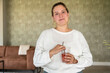 © Connect Images - Smiling woman in a white sweater holding a glass with a drink and straw, standing in a modern living room.