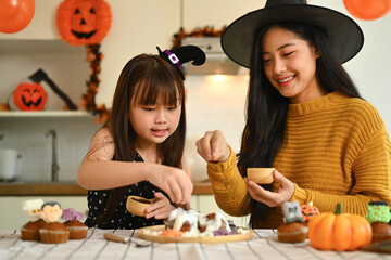 Beautiful young mother taking a picture of cute daughter with mobile phone decorating spooky cupcakes in kitchen