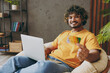 © ViDi Studio - Young IT man wear casual clothes work use laptop pc computer hold credit bank card shopping online sit in armchair stay home hotel flat rest relax spend free spare time in living room. Lounge concept.