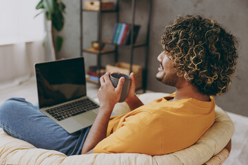 Wall Mural - Young Indian IT man wearing orange casual clothes hold use work on laptop pc computer drink tea sits in armchair stay at home hotel flat rest relax spend free spare time in living room Lounge concept