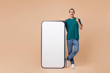 Wall Mural - Full body young middle eastern man wears blue t-shirt casual clothes big huge blank screen mobile cell phone smartphone with workspace area point finger up isolated on plain pastel beige background.