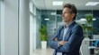 © lililia - A businessman in a suit stands with his arms crossed in a modern office lobby, with large windows and a polished floor