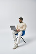 © LIGHTFIELD STUDIOS - A bearded man in a white sweater sits on a chair, engrossed in his laptop on a grey background in a studio setting.