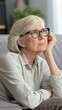 © lililia - A woman with grey hair wearing glasses and a shirt sits on a white sofa with a thoughtful expression. Her hand rests on a white pillow, while her other hand rests on her lap