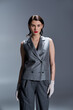 © LIGHTFIELD STUDIOS - Stylish young woman striking a pose in an elegant gray suit with a vest, accessorized with white gloves, in a studio on a grey background.