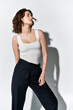 © LIGHTFIELD STUDIOS - A stylish young woman striking a pose in front of a white wall in a studio, wearing black pants and a white tank top.