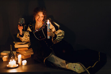 Wall Mural - Young adult woman dressed in a medieval dress sitting near ancient books and candles with bottle of red wine and holding a glass of wine in darkness