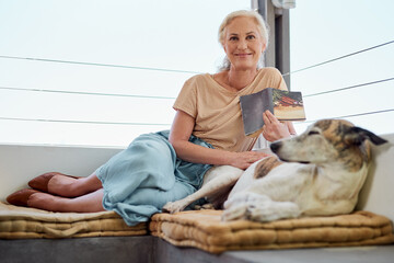 Poster - Elderly woman, reading book and portrait with dog on sofa for learning, knowledge or comfortable together on balcony. Animal, story and senior person with pet for hobby, literature or relax at home