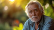 © RISHAD - Elderly man with gray hair and beard, wearing a denim shirt, looks thoughtfully into the distance in a green outdoor setting.