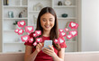 © Prostock-studio - A young chinese woman with long dark hair smiles as she uses her smartphone in a living room setting. Around her are pink hearts with white centers, simulating social media notifications.