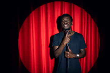 Wall Mural - Smiling African American man speaking to microphone performing in comedy show open mike stand up on stage with red curtains.