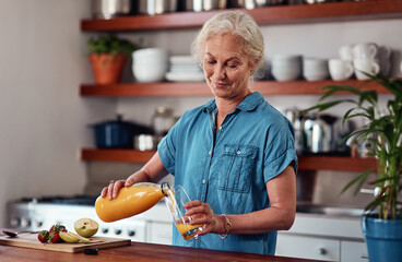 Glass, juice and senior woman at kitchen counter for breakfast, nutrition or detox treatment. Morning, bottle and elderly person pouring drink for vitamin C, fruit beverage or antioxidants in home