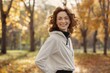 © Aaron Weiss - A woman with curly hair is smiling and wearing a white hoodie. She is standing in a park with trees and leaves on the ground
