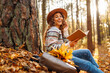 © maxbelchenko - Beautiful smiling young woman student sitting and reading book at autumn park fall yellow foliage leaves. Concept of enjoying nature.