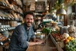 © Vera - Man sitting at a table in a coffee shop, smiling at the camera while working on his laptop. The atmosphere is cozy and inviting, portraying a successful small business owner