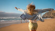© Andrii - Carefree woman in denim shirt with blond hair running on sandy beach. Back view of cheerful lady with arms outstretched enjoying summer vacation along sea against blue sky.