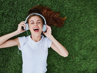 Wall Mural - A girl listening to music in headphones lying on the green grass in the park and smiling in a white T-shirt, summer mood as a lifestyle