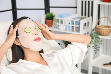 Sticker - Young Asian woman with cucumber slices and facial sheet mask relaxing in spa salon, closeup