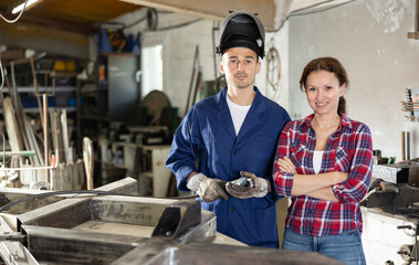 Wall Mural - Man and woman interact while working in workshop. Man trains woman to do connecting iron rails with welding machine. Two persons standing in storeroom, holding welding machine, resting after work