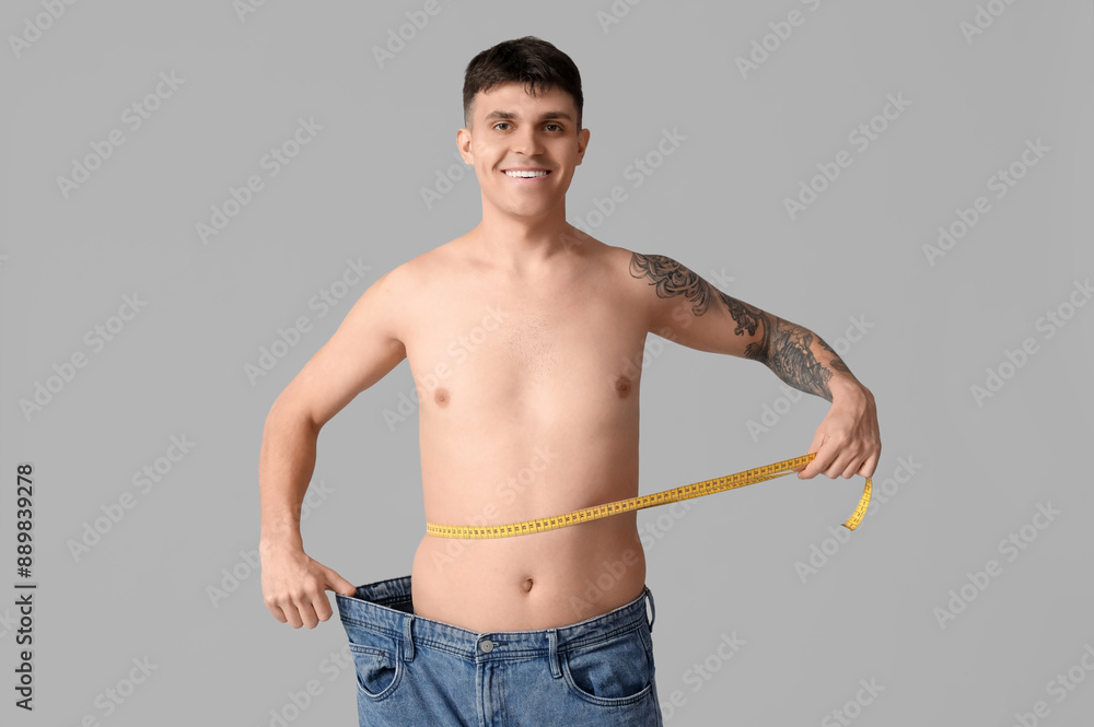 Young man in loose jeans and with measuring tape on grey background. Weight loss concept
