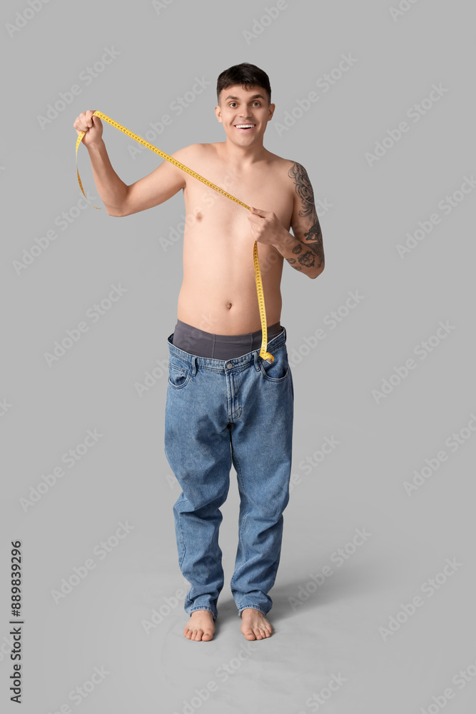 Young man in loose jeans and with measuring tape on grey background. Weight loss concept