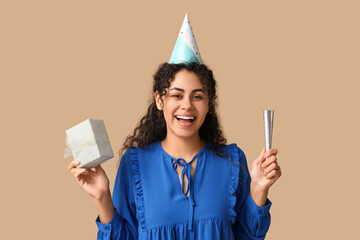 Poster - Beautiful young African-American woman in party hat with whistle and gift box on brown background