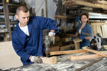 Male employee of carpentry workshop uses drill to make holes in wooden billet, drill surface, prepare wooden products for further assembly and installation