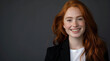 © GreenOptix - attractive female red head smiling, wearing black blazer and white tshirt on the right side of frame, long hair with soft waves falling over shoulders, standing in front of dark grey background