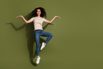 Full size photo of pretty girl dressed beige t-shirt jeans jumping arms presenting objects empty space isolated on khaki color background