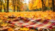 © Musarrof - A plaid blanket spread on the ground amidst fallen autumn leaves in a park, capturing a cozy and inviting fall setting.