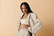 © LIGHTFIELD STUDIOS - A young woman with long braids poses confidently in a beige blazer and pants, looking down thoughtfully against a neutral backdrop.