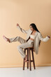 © LIGHTFIELD STUDIOS - A young woman wearing a beige suit sits on a wooden stool, one leg extended, in a studio setting.