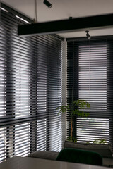 Leinwandbilder - Wooden automatic blinds partially open, covering large corner windows in the interior. Sunlight gently filters through wood venetian blinds, creating a serene atmosphere, complemented by indoor plants