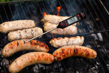 Close-up shot of digital meat thermometer with Celsius scale showing safe cooking temperature inside of grilling sausages on smoking barbecue