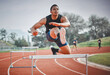 © DavisShared/peopleimages.com - Hurdles, fitness and man running on track at stadium training for race, competition or marathon. Energy, jump and male athlete sprint by equipment for cardio workout or exercise at sports arena.
