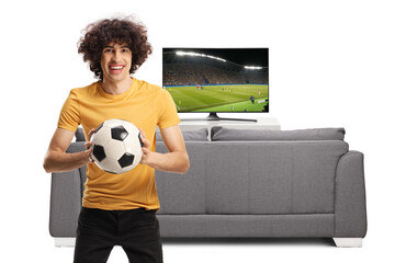 Wall Mural - Young passionate football fan holding a ball and smiling at camera in front of tv and sofa