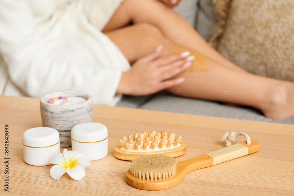 Table with spa composition and young woman applying body scrub on legs at home