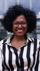 Wall Mural - Vertical. Camera zoom of adult African American business woman posing smiling standing with hands clasped together in front office building. Cheerful black female looking happy at camera outdoors