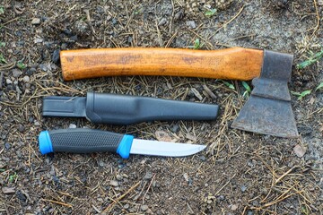 one with a black handle and a plastic cover, a sharp iron shiny dangerous knife with a small industrial old rusty hatchet with a wooden handle lying on the ground during the day on the street