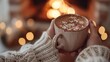 © Onzdemia - Close-up of hands holding a warm mug of coffee or hot chocolate in front of a cozy fireplace.