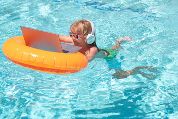 Wall Mural - Child sitting in swimming ring in pool and using laptop. Shopping online, freelance concept, summer travel.