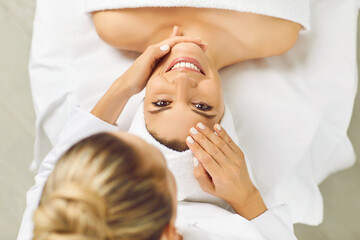 Wall Mural - Top view portrait of happy smiling young pretty woman client looking cheerful at camera while lying in spa salon having a face massage and receiving beauty procedure from beautician in beauty clinic.