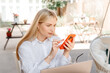 © Anton Pentegov - young beautiful blond caucasian woman sitting at table in outdoor terrace of cafe in summer day with notebook, businesswoman working, confident freelancer watching smartphone
