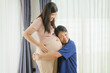 © NanSan - An Asian husband sits on a sofa, gently holding his pregnant wife's stomach. He lovingly cares for her as they eagerly await the arrival of their first child, sharing a tender moment.