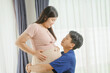 © NanSan - An Asian husband sits on a sofa, gently holding his pregnant wife's stomach. He lovingly cares for her as they eagerly await the arrival of their first child, sharing a tender moment.
