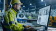 © VK Studio - A construction engineer wearing safety gear is focused on designing structural plans on a computer in a busy industrial environment.