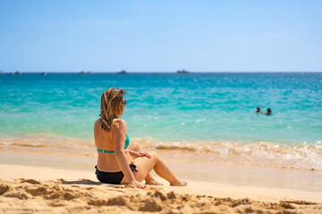 Wall Mural - Beach vacation. Happy young woman in bikini sitting on sandy beach by the sea on beautiful sunny day. Positive emotion
