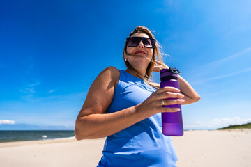 Drinking water in hot summer. Portrait of young modern woman with long blonde hair drinking water from water bottle on sandy beach on sunny day. Break from sports training to replenish water in body