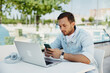 © SHOTPRIME STUDIO - Young man working remotely on laptop, checking smartphone, outdoor café setting, focused and relaxed