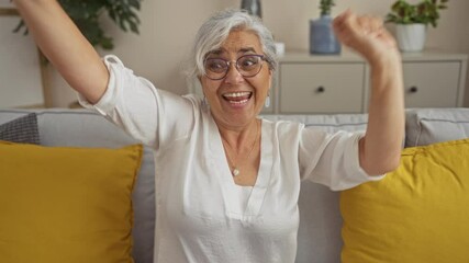 Wall Mural - Elderly woman with grey hair and glasses joyfully dancing on a cozy living room sofa with yellow pillows and indoor plants.